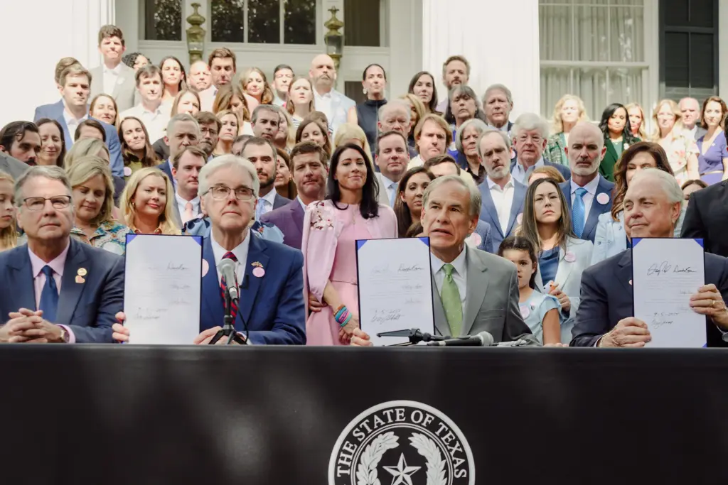 Gov. Greg Abbott, flanked by state Sen. Charles Perry, Lt. Gov. Dan Patrick and state Rep. Drew Darby, and backed by families of the Camp Mystic flood victims known as the “Heaven’s 27,” holds up the signed Senate Bill 1 and House Bill 1 at the Governor’s Mansion on Sept. 5, 2025. The bills, passed during the second special session of the 89th Texas Legislature, imposed stringent fees and requirements on camps.