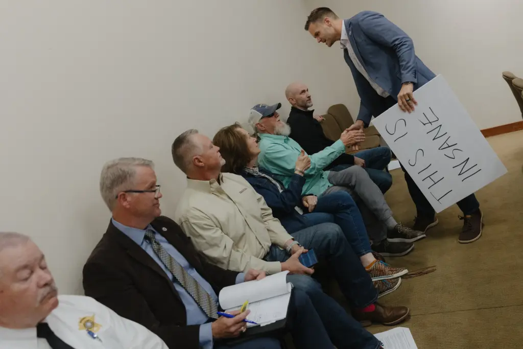 Matt Long shakes hands with attendees while holding a sign that says “This is insane” of a Hood County Commissioners Court meeting in Granbury on February 23, 2026. The pause would allow for the county to assess water and power usage of the proposed eight data centers that would be built in the county.
