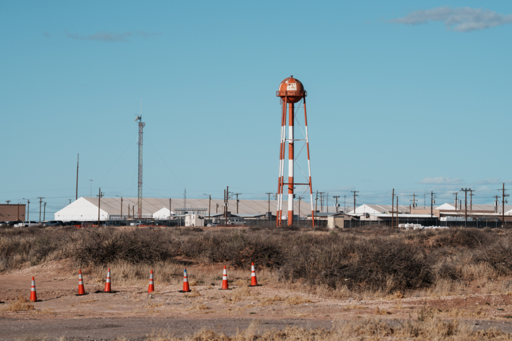 A row of orange traffic cones lines a dry, scrubby dirt field in the foreground. In the background, long, white tent-like buildings and a prominent orange-and-white striped water tower stand under a clear blue sky.